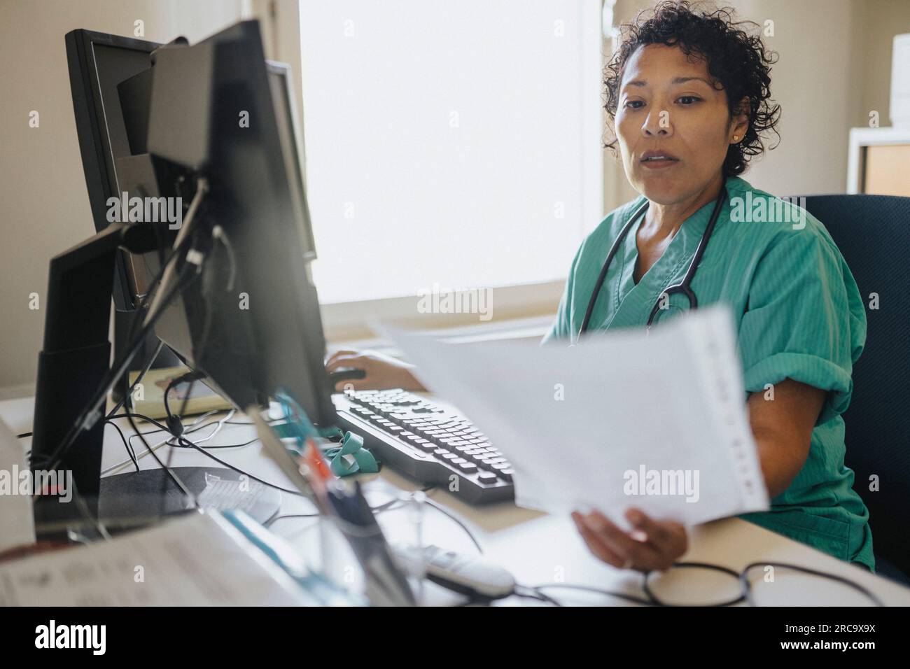 Medico femminile che esamina la cartella clinica mentre si utilizza il computer alla scrivania in ufficio Foto Stock