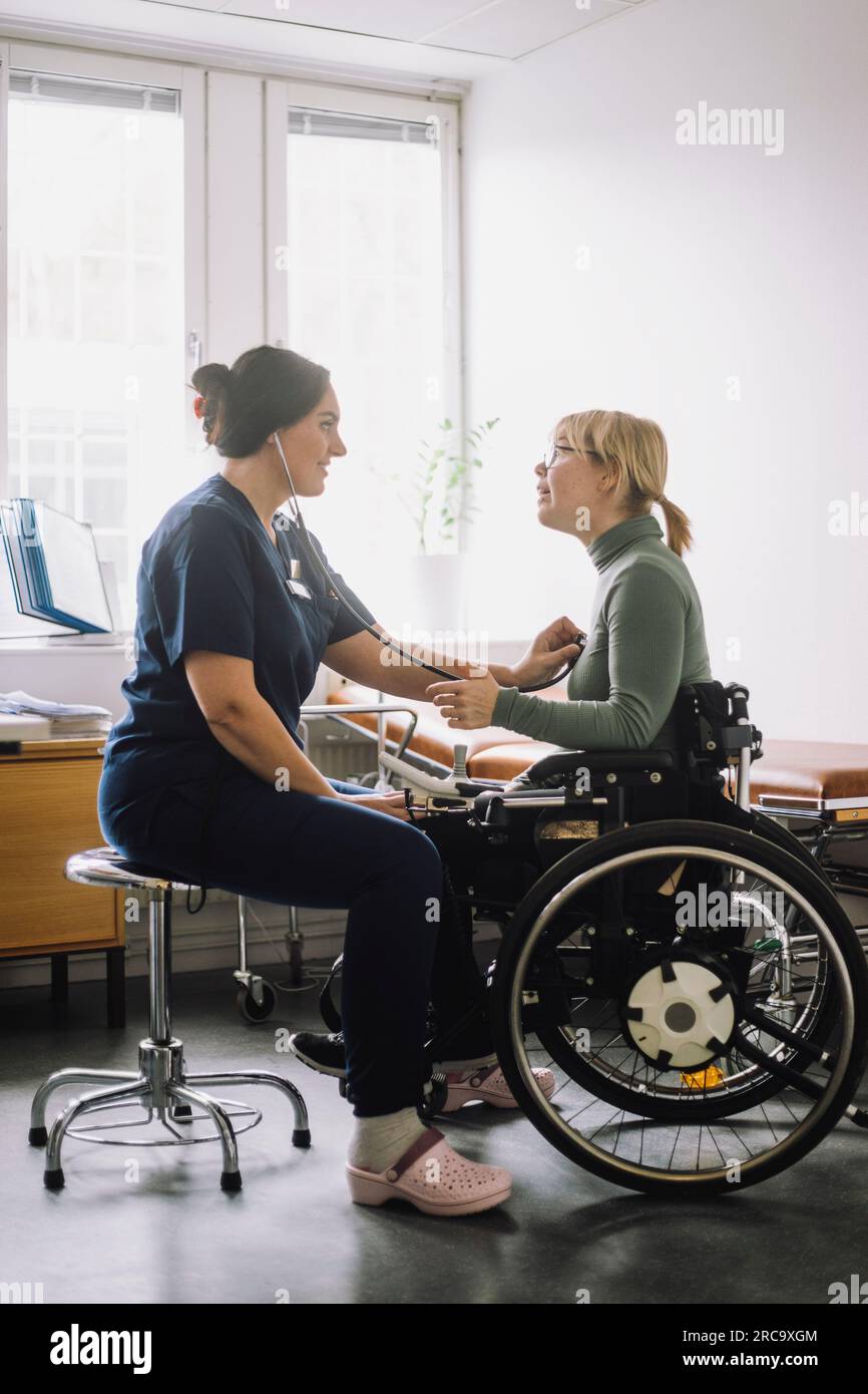 Lavoratore sanitario femminile che ascolta il battito cardiaco del paziente seduto su sedia a rotelle in clinica Foto Stock