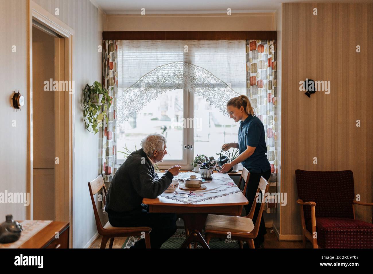 Uomo anziano che parla con una badante che annaffia le piante a casa Foto Stock