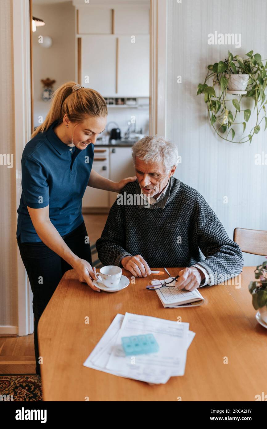 Assistente di cura femminile che serve tè a un anziano seduto a casa Foto Stock