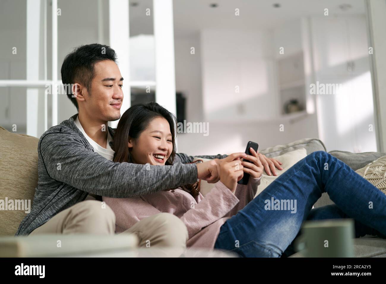 felice giovane coppia asiatica seduta sul divano della famiglia a casa guardando insieme le foto del cellulare Foto Stock