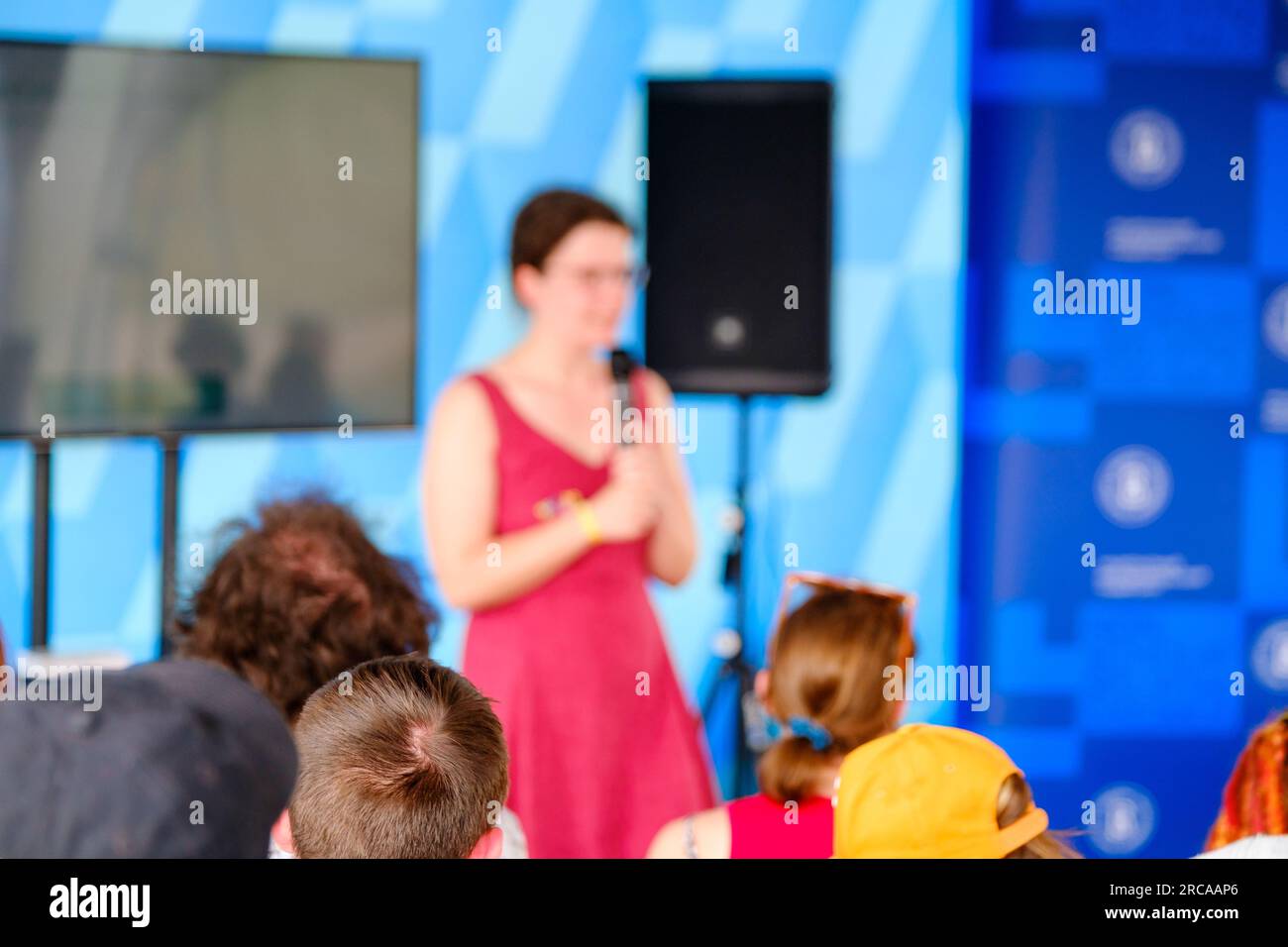 Vista posteriore di persone irriconoscibili sedute sulle sedie e ascoltando una speaker femminile anonima in piedi sul palco con microfono vicino al monitor durante la fase B. Foto Stock