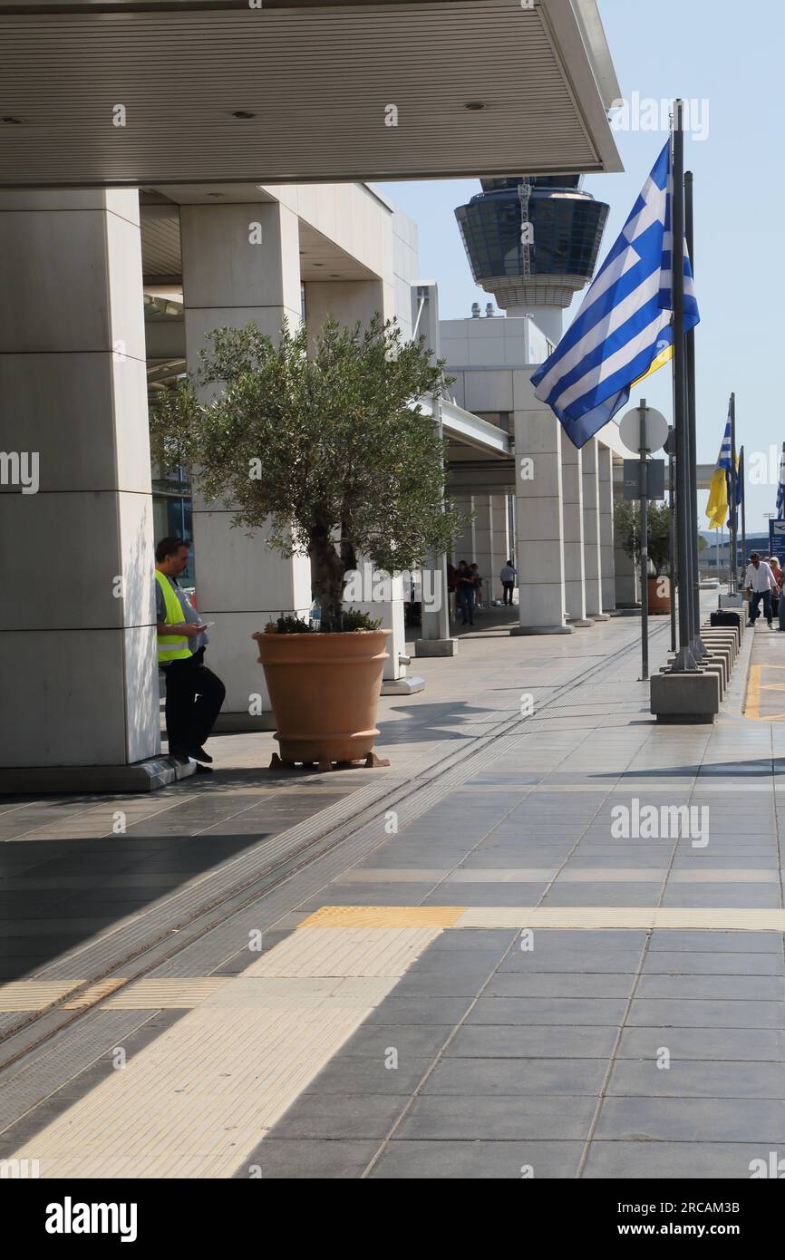 Atene Grecia Aeroporto Internazionale di Atene (AIA) Eleftherios Venizelos Man Wearing High Visibility Gilet Standing Olive Tree in Pot Outside Foto Stock