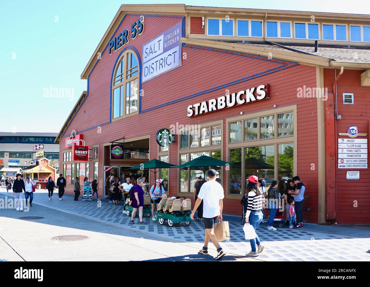 Persone che camminano lungo il lungomare pedonale sotto il forte sole pomeridiano con uno Starbucks al Pier 55 Seattle, Washington State USA Foto Stock