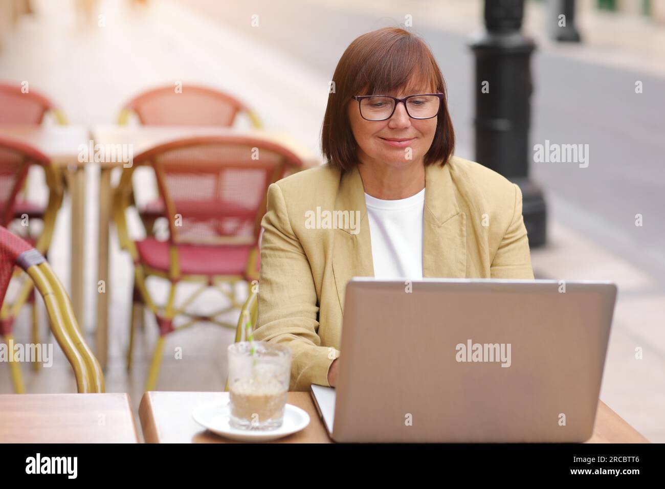 Donna d'affari matura ed elegante con occhiali da vista lavora sul computer portatile mentre si siede con un caffè all'aperto in un caffè europeo. Concetto di lavoro remoto da pub Foto Stock