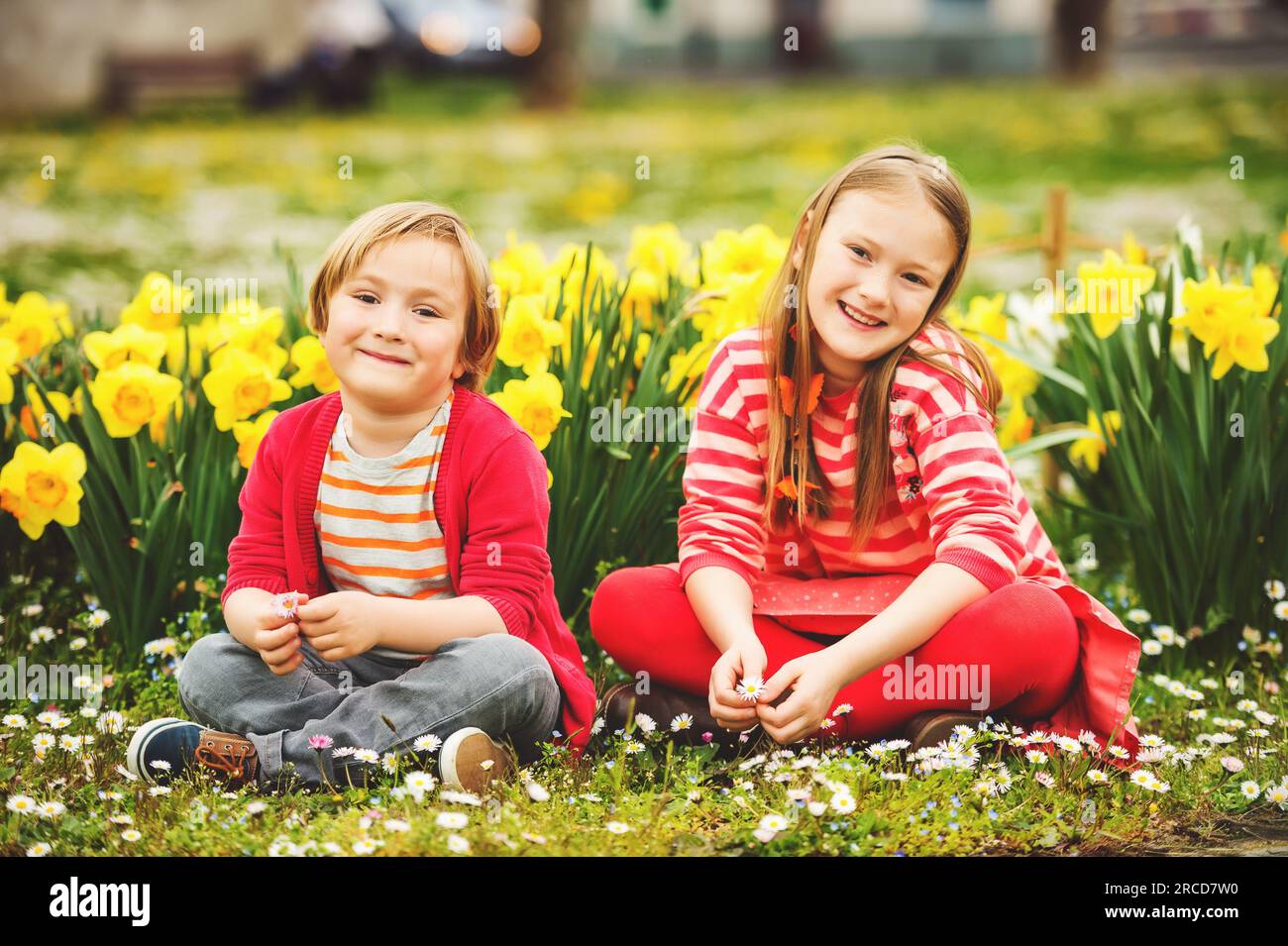 Due bambini carini, il bambino e la sorella maggiore, che giocano nel parco tra i fiori di narcisi gialli, indossando abiti rossi accesi Foto Stock