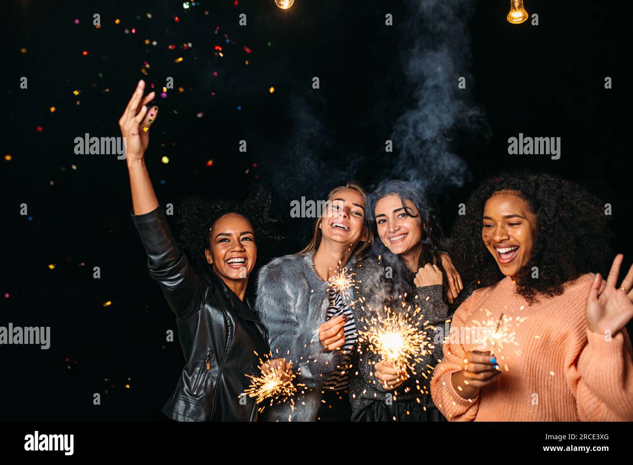 Quattro donne diverse che si divertono all'aria aperta tenendo scintille e lanciando coriandoli Foto Stock