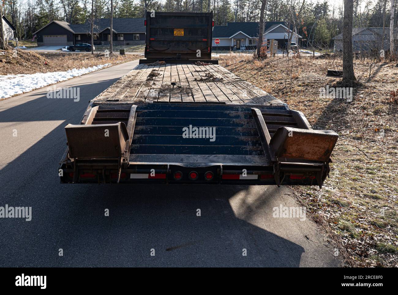 Il rimorchio a pianale vuoto con rampe rialzate è parcheggiato sulla strada asfaltata in un quartiere con case in lontananza, al sole nel tardo pomeriggio. Foto Stock