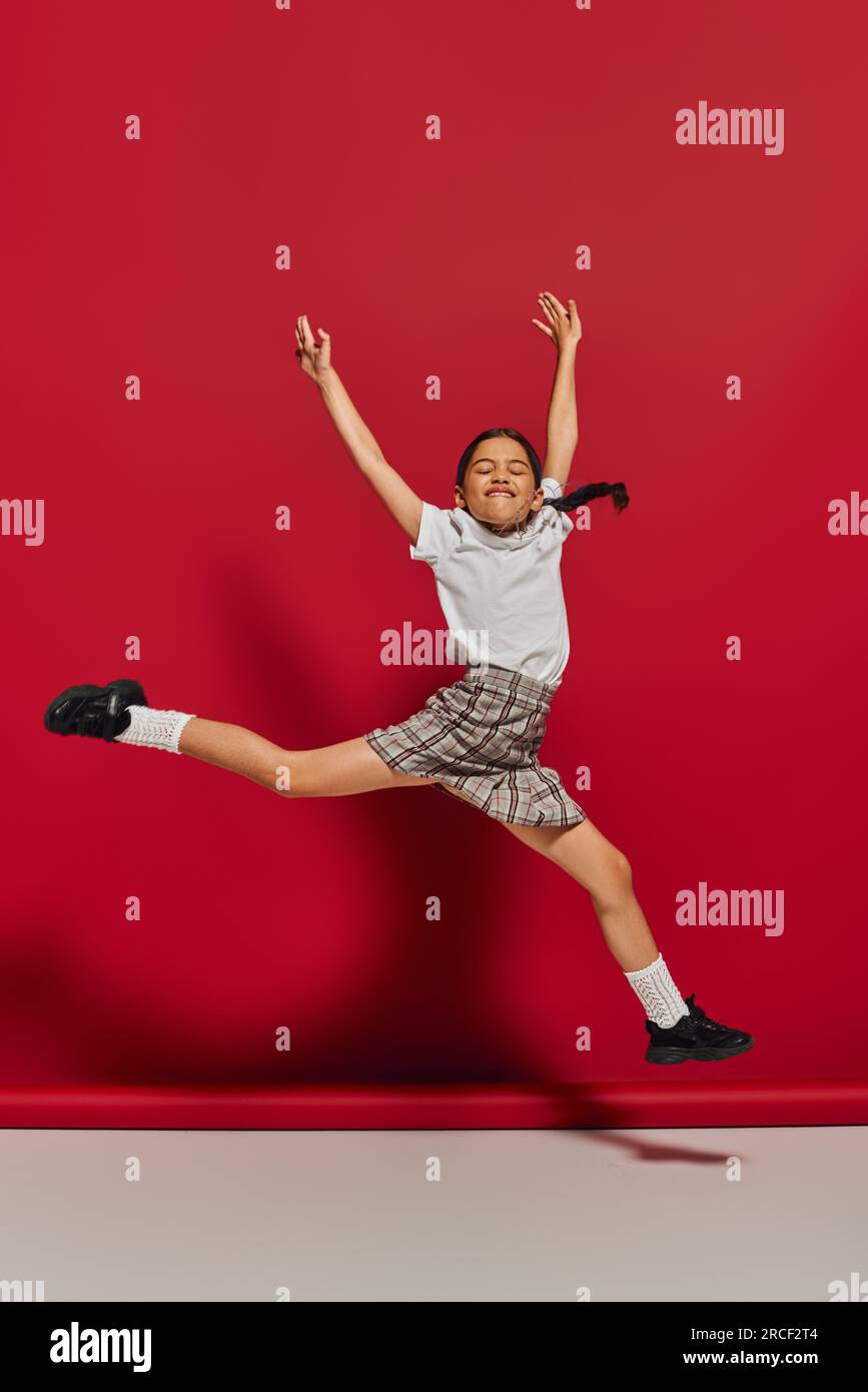 Eccitata e allegra ragazza preadolescente con t-shirt bianca ed elegante gonna a scacchi che salta e si diverte mentre posa su sfondo rosso, acconciatura e tr Foto Stock