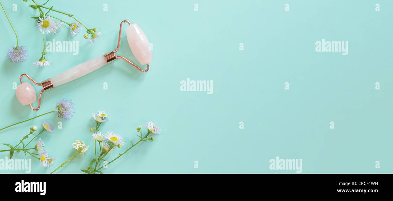Rullo massaggiante per il viso in quarzo rosa su sfondo blu con fiori. Massaggiatore facciale antirughe e anti-invecchiamento, strumento per la cura della pelle Foto Stock