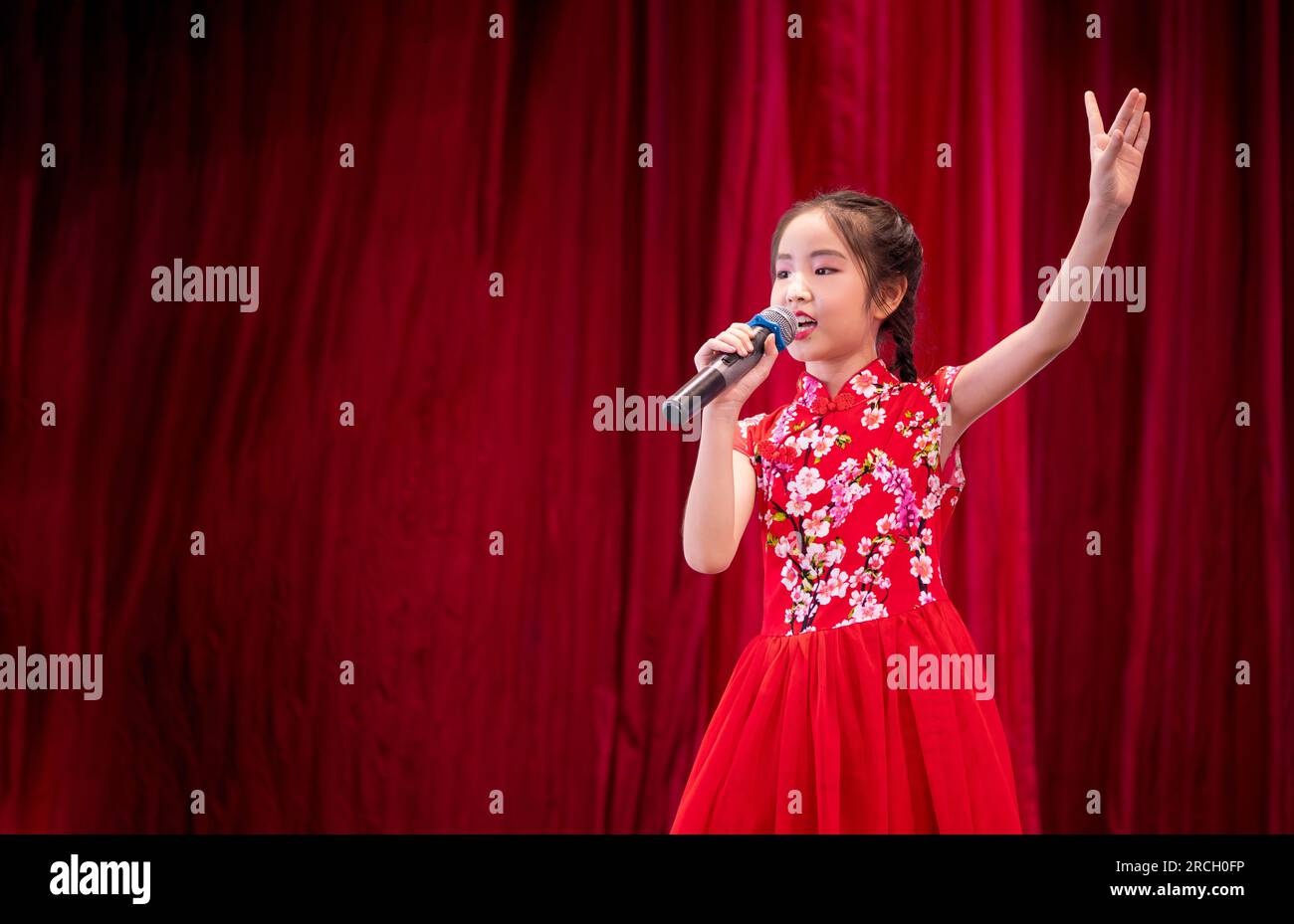 La bambina asiatica canta una canzone sul palco durante la giornata di attività scolastica, vestita in stile Qipao, sfondo rosso tenda Foto Stock