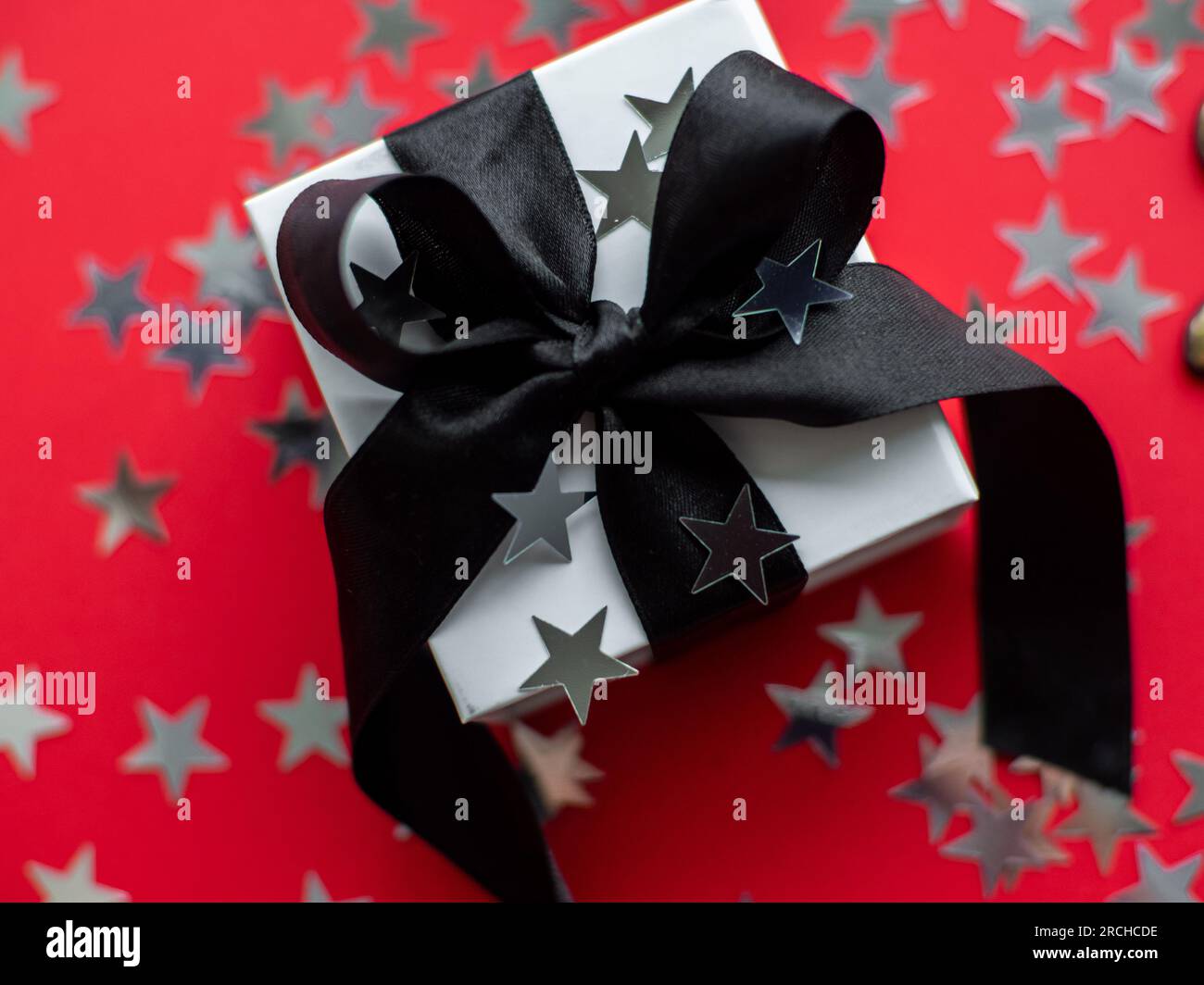 Sfondo rosso con confezione regalo avvolta con nastro nero e coriandoli a forma di stelle argentate. Il concetto di biglietto d'auguri per il nuovo anno. Natale festivo Foto Stock