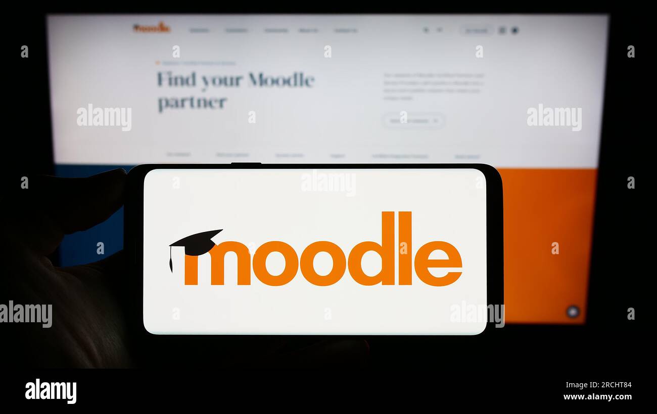 Persona che possiede un telefono cellulare con il logo della piattaforma di apprendimento Moodle sullo schermo davanti alla pagina Web. Concentrarsi sul display del telefono. Foto Stock