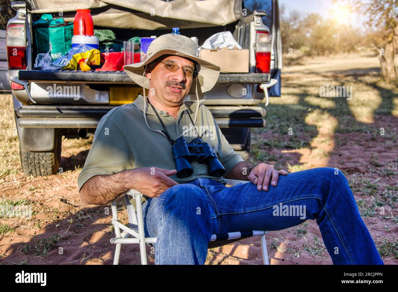 uomo arabo con binocolo e cappello da safari seduto su una sedia di fronte al suo veicolo 4x4 carico che ha una pausa barbecue Foto Stock