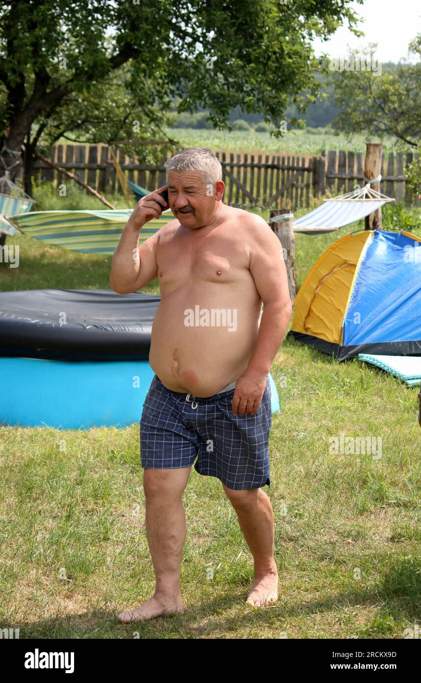 Uomo Mustachioed in pantaloncini. Parla al telefono. Prato all'aperto. Vicino alla piscina e alle amache. Foto Stock