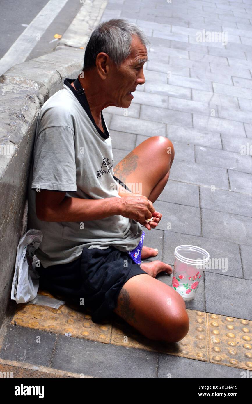 Un uomo anziano o anziano siede su un marciapiede nel centro di Bangkok, Thailandia, Asia; di fronte a lui c'è una coppa per invitare donazioni; conta le sue monete Foto Stock