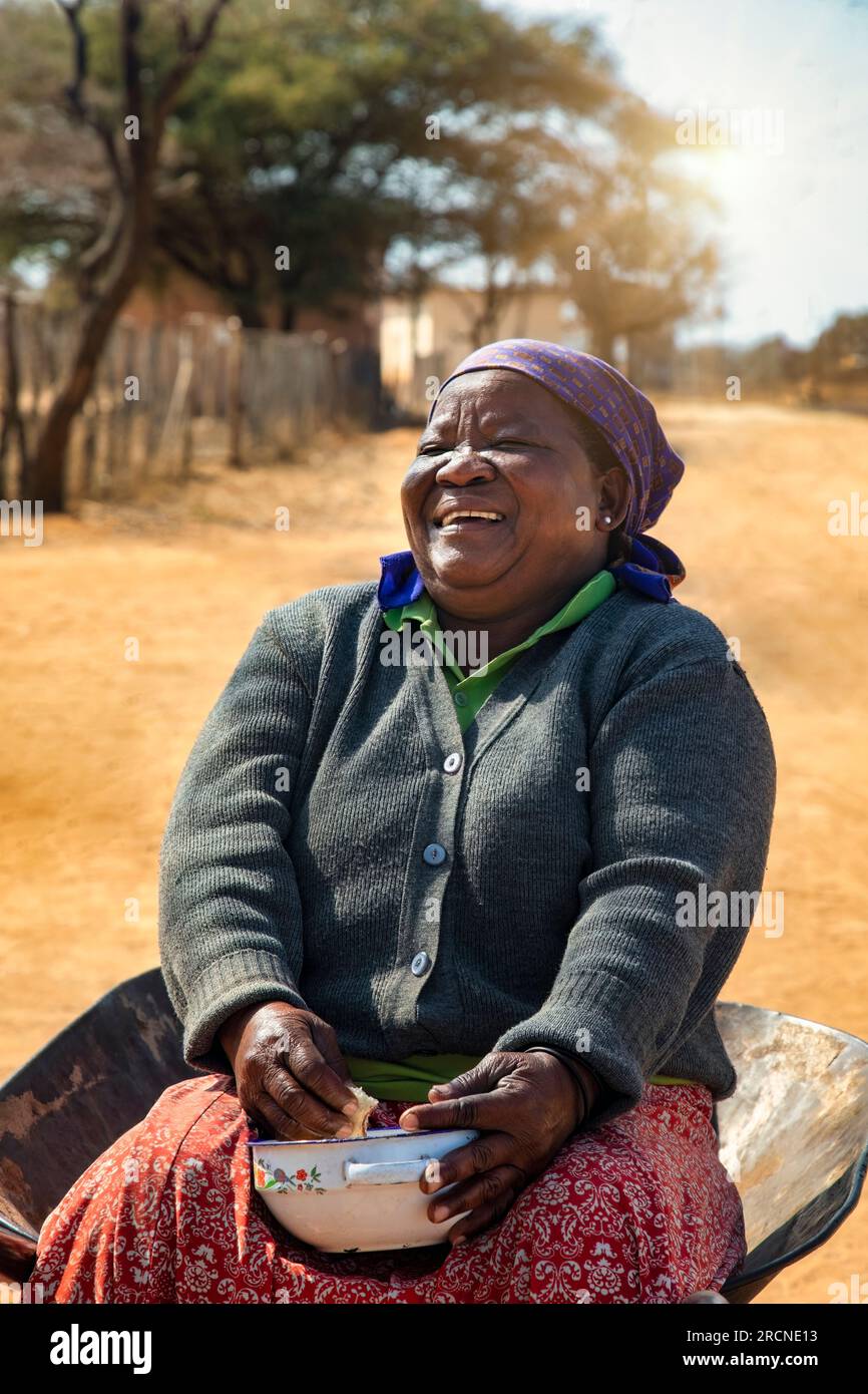 vita da villaggio, vecchia donna africana seduta in una carriola a mangiare stufato e ridere dal cuore Foto Stock