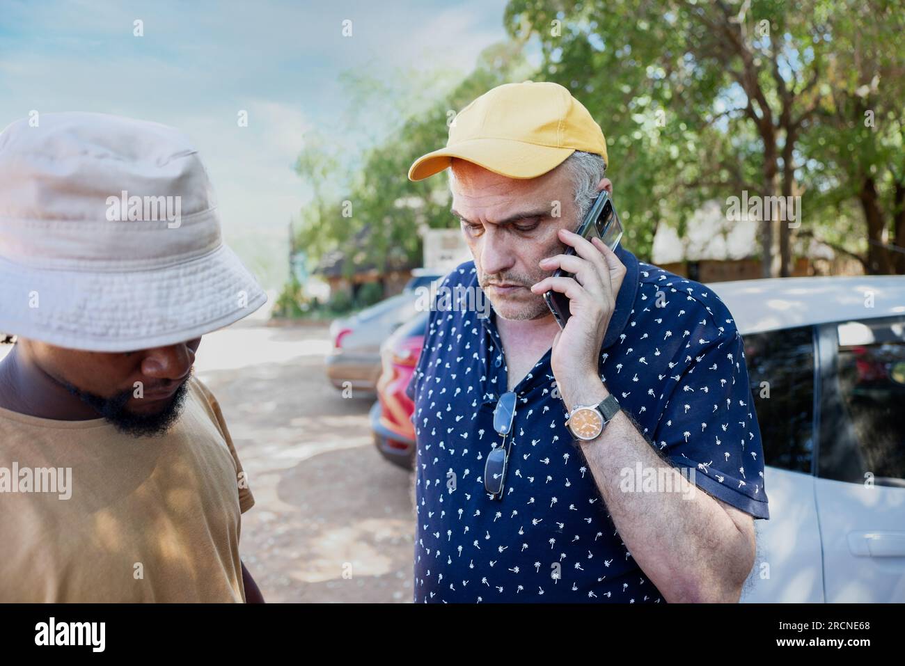 vecchio con un berretto giallo, che parla al telefono, il suo giovane amico africano nelle vicinanze, in piedi su una strada trafficata di fronte a poche auto Foto Stock