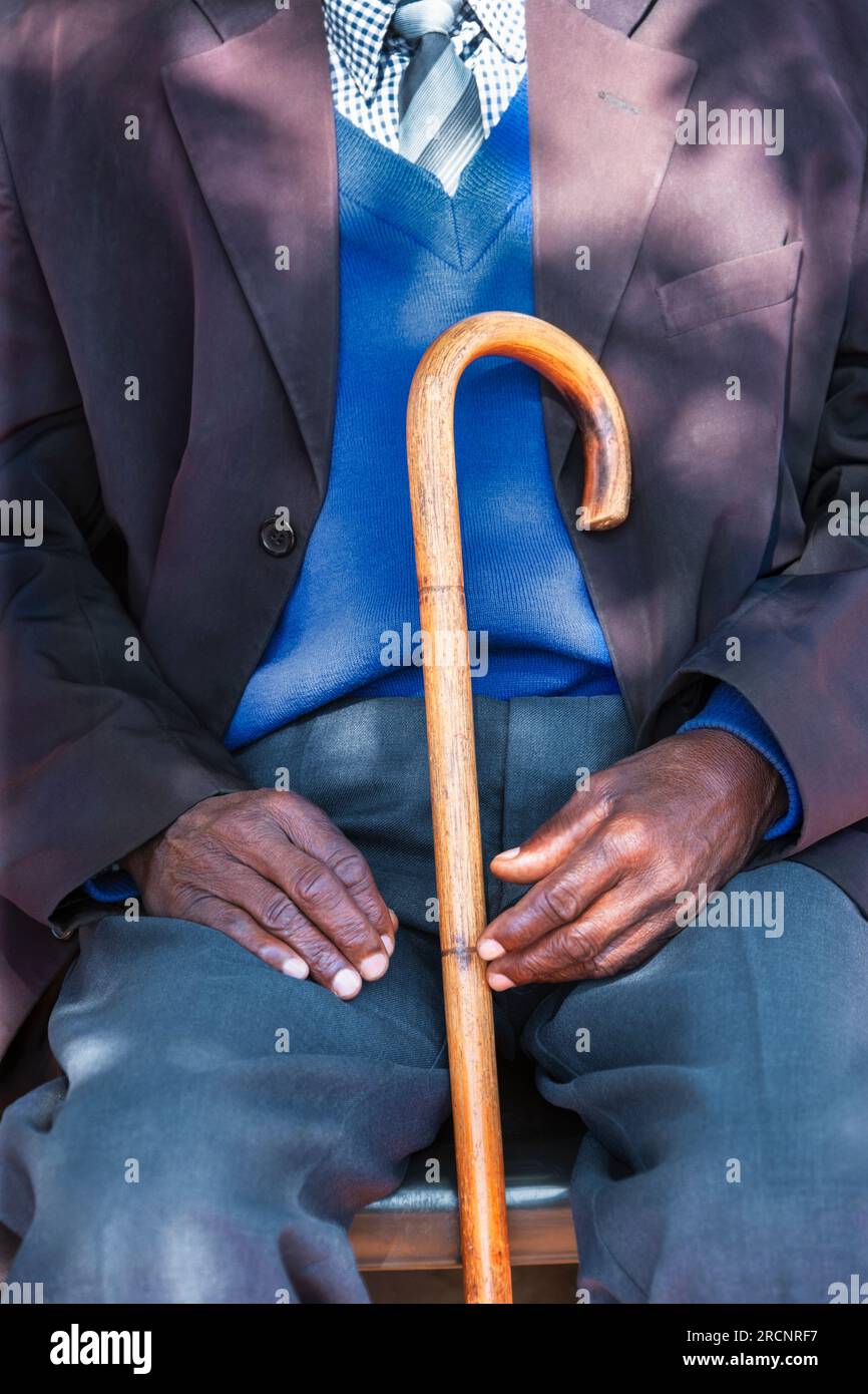 vecchio africano stanco ed elegante con un bastoncino che si appoggia su una sedia. all'aperto nel cortile del suo villaggio Foto Stock