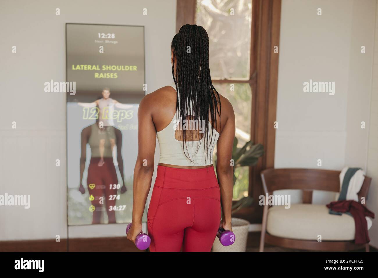 Con l'aiuto di un'istruttrice virtuale su uno specchio per il fitness intelligente, una donna si impegna in esercizi di sollevamento pesi, utilizzando la tecnologia avanzata per il fitness di ge Foto Stock