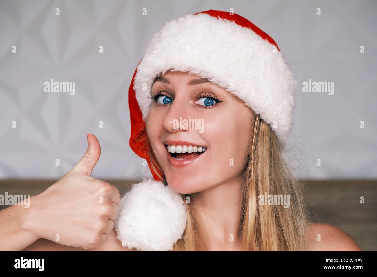 Sorridente Babbo Natale che indossa un cappello rosso che gestisce i pollici verso l'alto guardando la fotocamera. Ritratto di una giovane donna bionda dagli occhi blu con il cappello di Babbo Natale. Natale Foto Stock