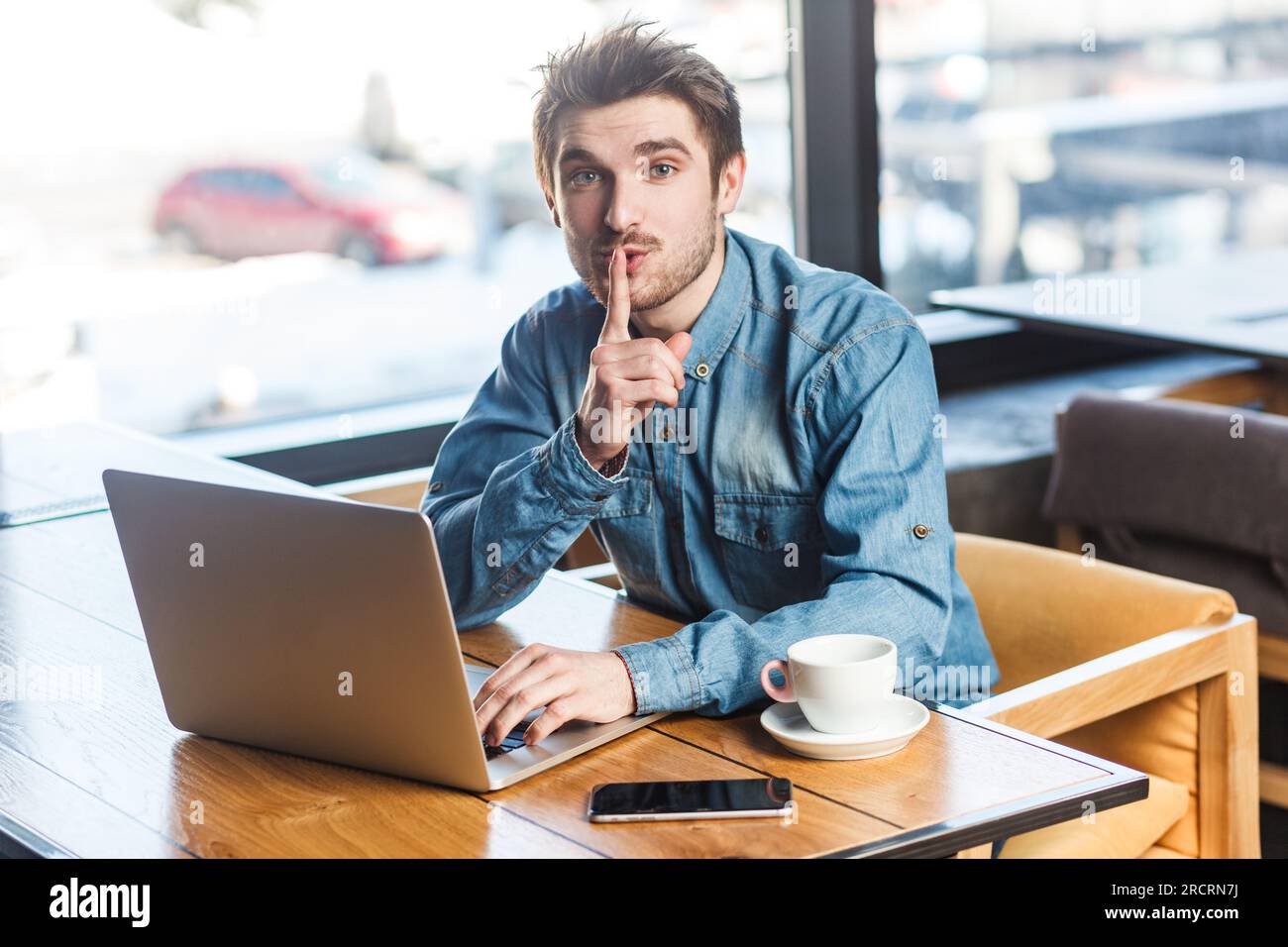 Ritratto di un bel giovane uomo barbuto freelance in camicia blu jeans che lavora su un notebook, mostra un gesto shh, guarda la fotocamera, chiede di non fare rumore. Foto al coperto vicino a una grande finestra, sfondo del caffè. Foto Stock