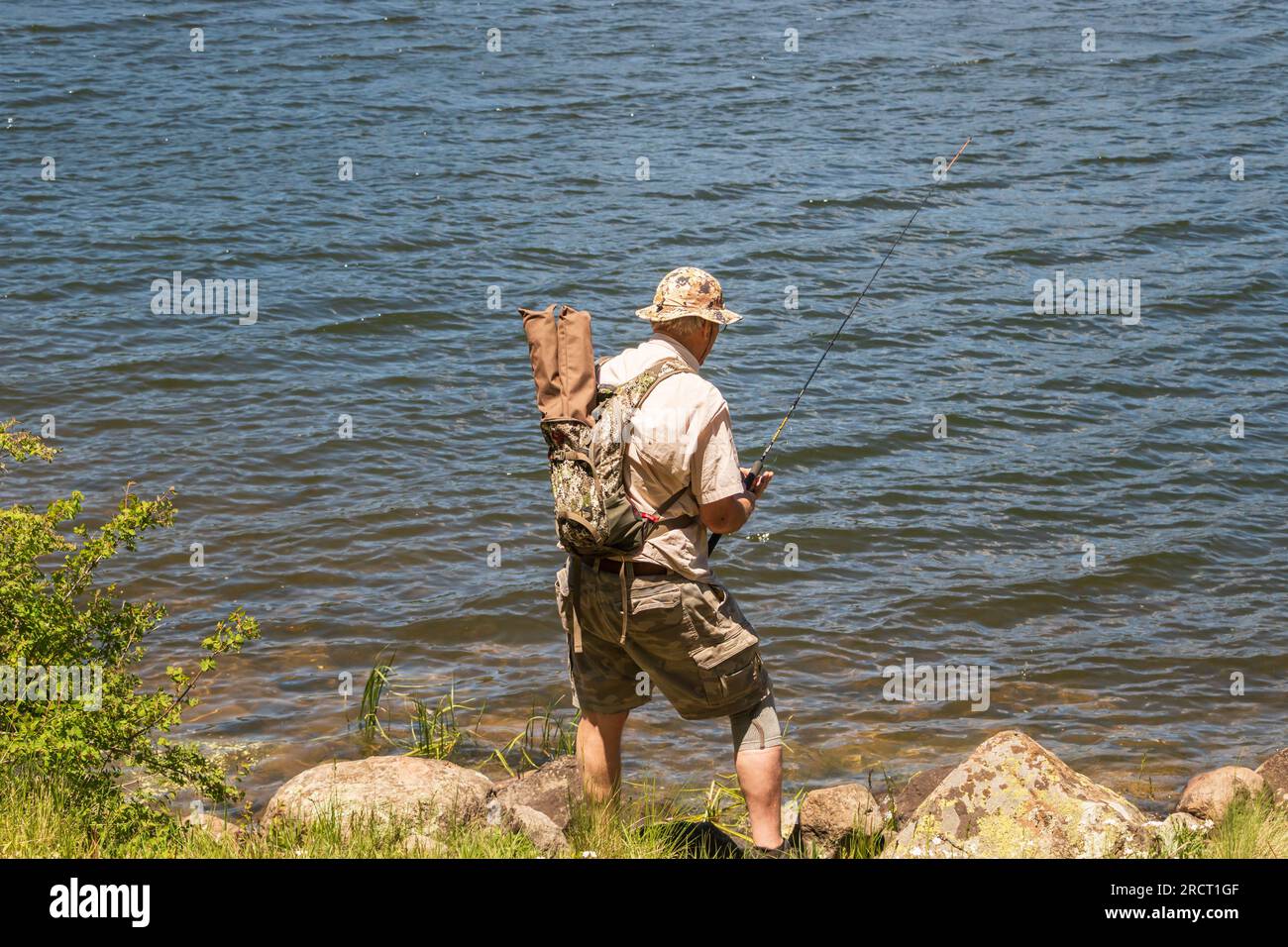 Un uomo anziano di età superiore ai 70 anni si trova accanto a un lago per la pesca con un ginocchio indossando uno zaino Foto Stock