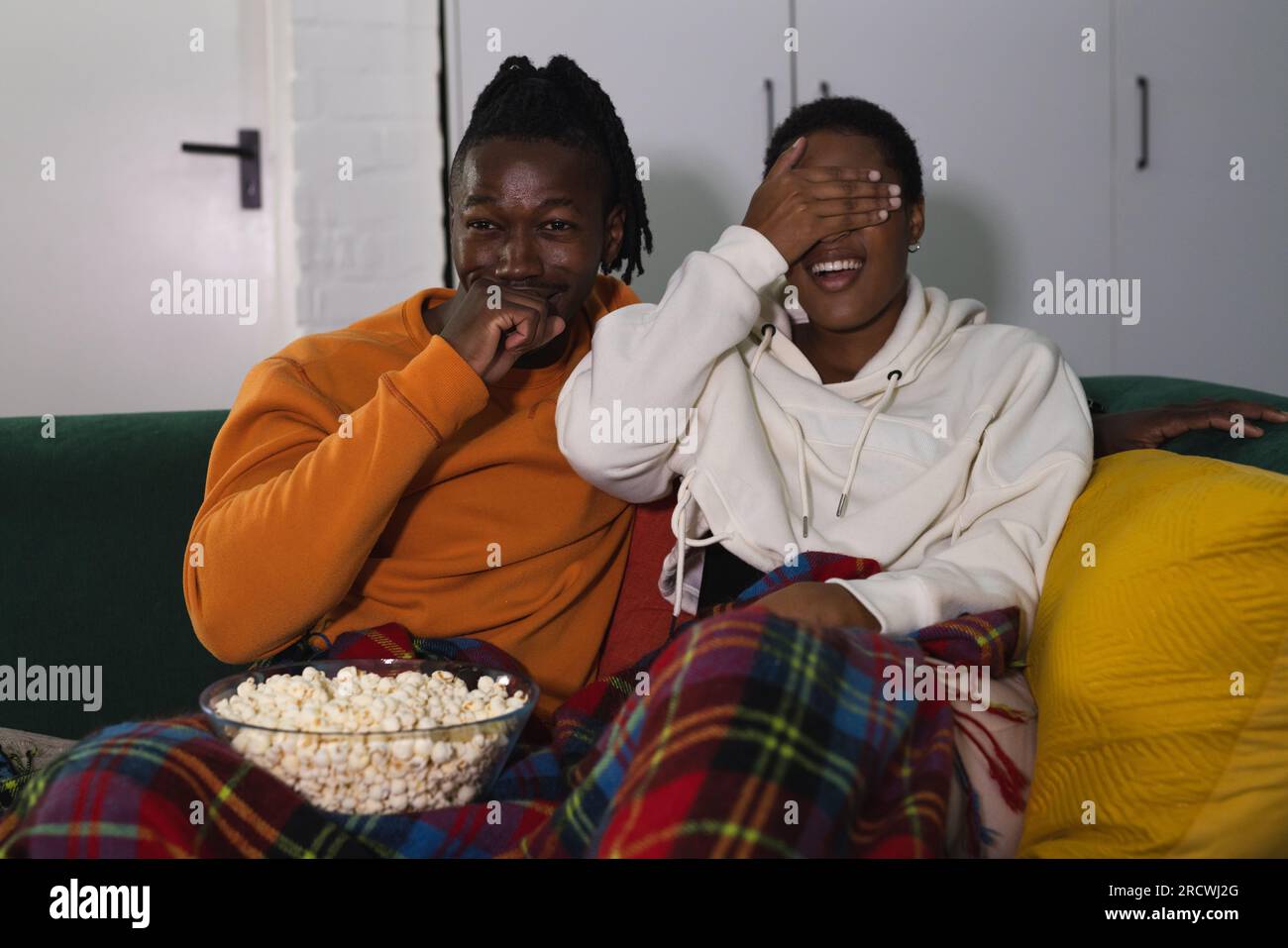 Coppia afro-americana spaventata che si rilassa a casa, copre gli occhi e guarda un film spaventoso con popcorn Foto Stock