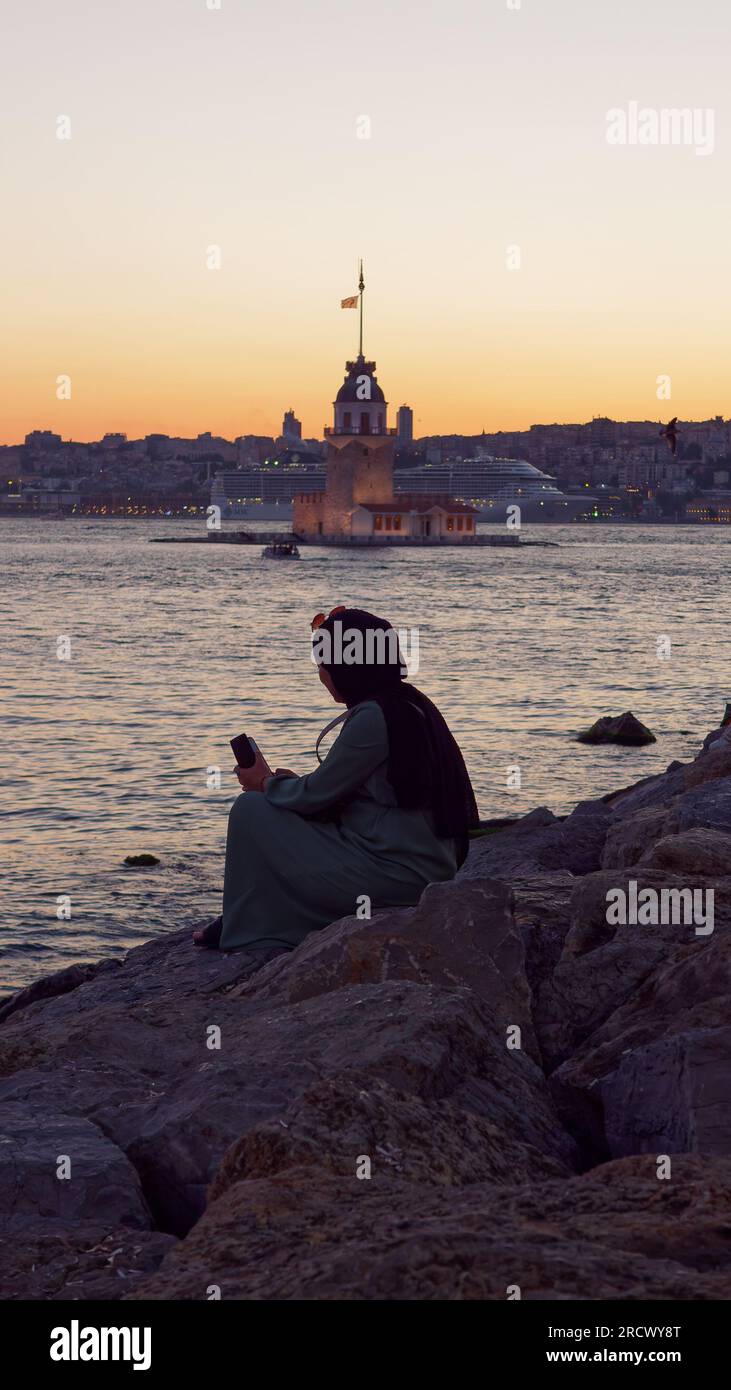 Signora musulmana con telefono e occhiali da sole siede sulle rocce e gode del tramonto sul Mare del Bosforo e sulla Torre delle Fanciulle da Uskudar, Istanbul, Turchia Foto Stock