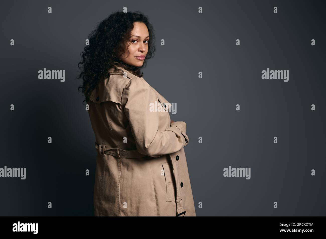 Ritratto in studio sicuro di una bella donna incinta dai capelli ricci latinoamericani, che indossa un elegante trench beige e guarda la fotocamera attraverso di lei Foto Stock