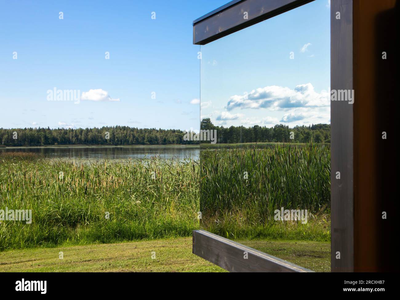 Vista dettagliata della moderna finestra riflettente a specchio sull'edificio nel mezzo della natura selvaggia. Cottage di lusso. Lago, canne, alberi, cielo blu. Foto Stock