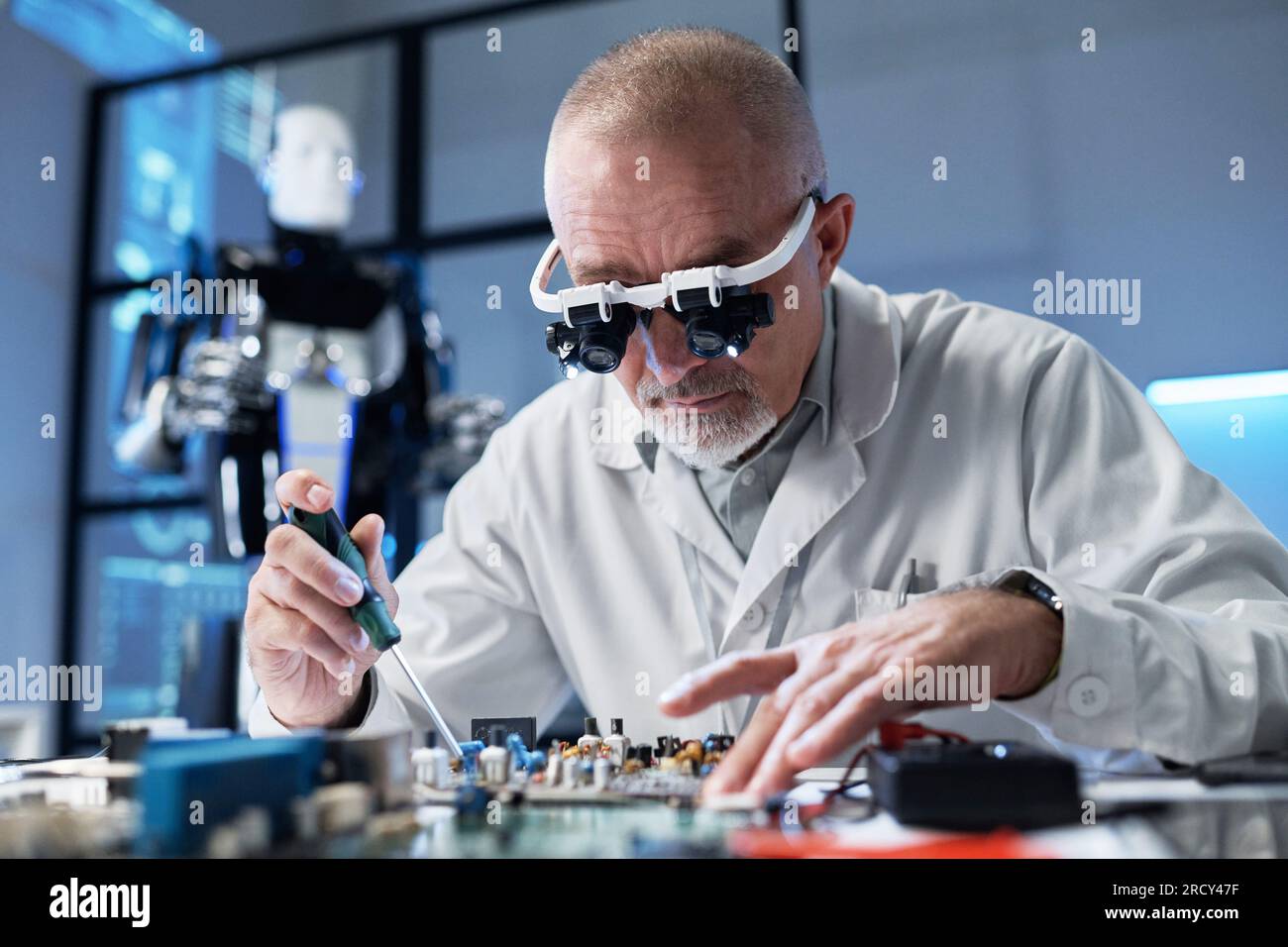 Programmatore maturo in camice da laboratorio seduto al tavolo e assemblato la scheda madre in ufficio Foto Stock