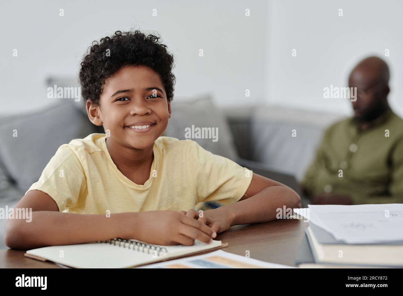 Ritratto di un ragazzo afro-americano sorridente alla macchina fotografica mentre sedeva al tavolo e faceva i compiti Foto Stock