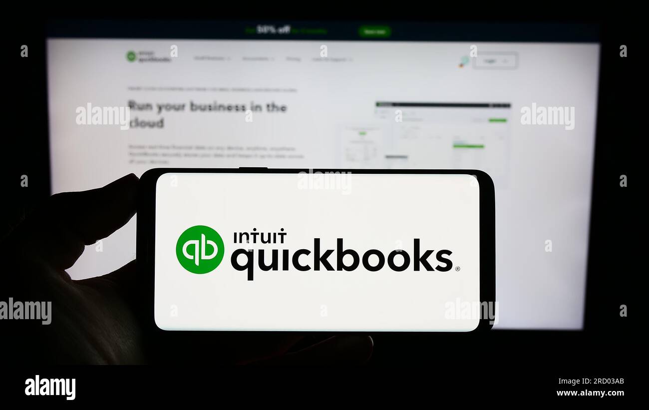 Persona che possiede uno smartphone con il logo del software di contabilità QuickBooks sullo schermo di fronte al sito Web. Concentrarsi sul display del telefono. Foto Stock