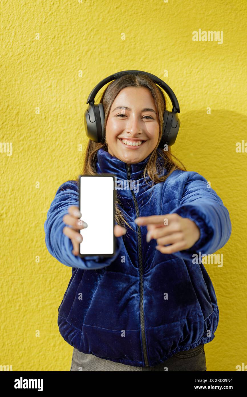 La ragazza latina con le cuffie mostra lo schermo vuoto del suo telefono cellulare isolato su uno sfondo giallo. Foto Stock