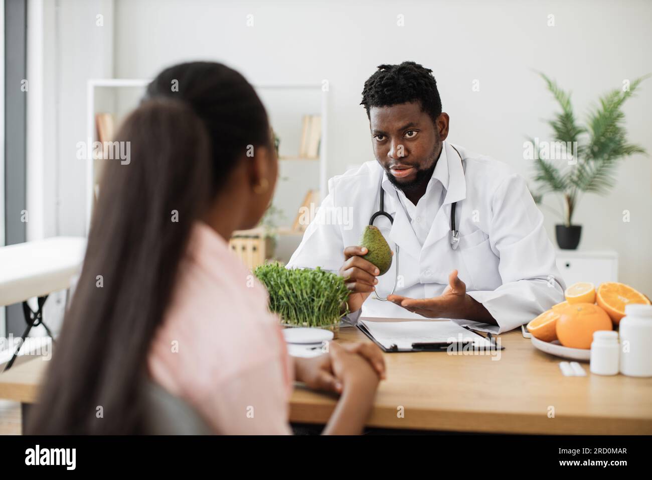 Uomo afro-americano concentrato in un cappotto bianco che tiene avocado mentre parla con una cliente femminile in una sala di consulenza. Specialista medico in cibo incoraggiando ad arricchire mangiare con frutta piena di nutrienti. Foto Stock