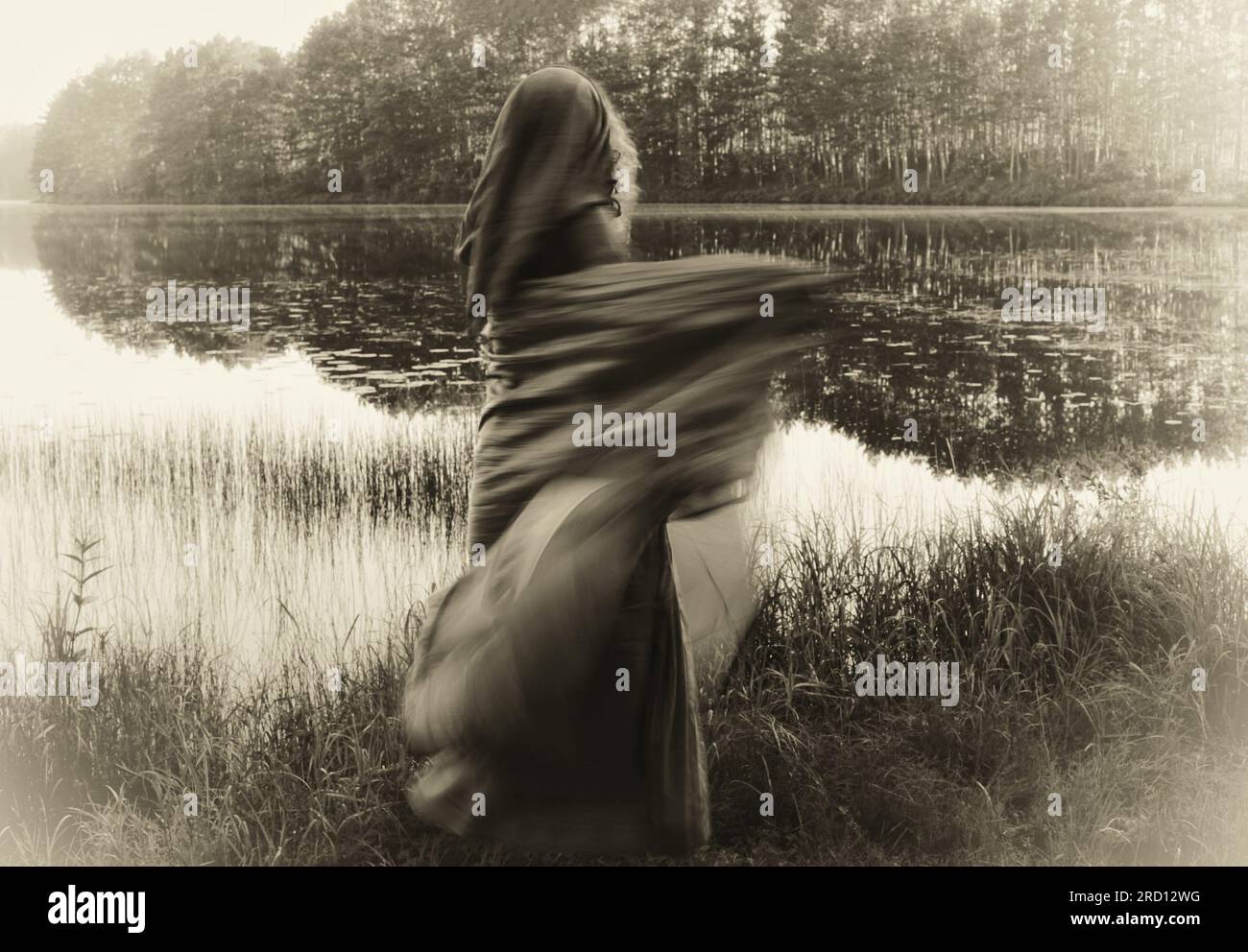 Sciamana in abito con cappuccio, danzante in movimento sfocato. Saluta la mattina di un lago magico. La foto evoca una qualità fantasma ultraterrena. Foto Stock