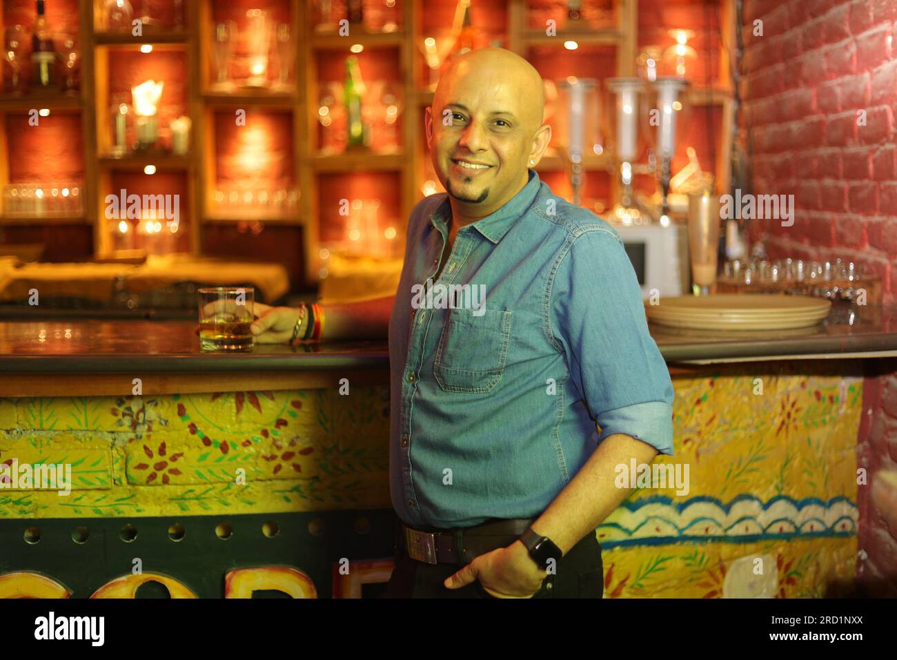 Scena drammatica di un giovane e sfavillante ragazzo calvo in piedi sul bancone del bar. Goditi l'interno del club cittadino mentre il night club regge un bicchiere di whisky Foto Stock