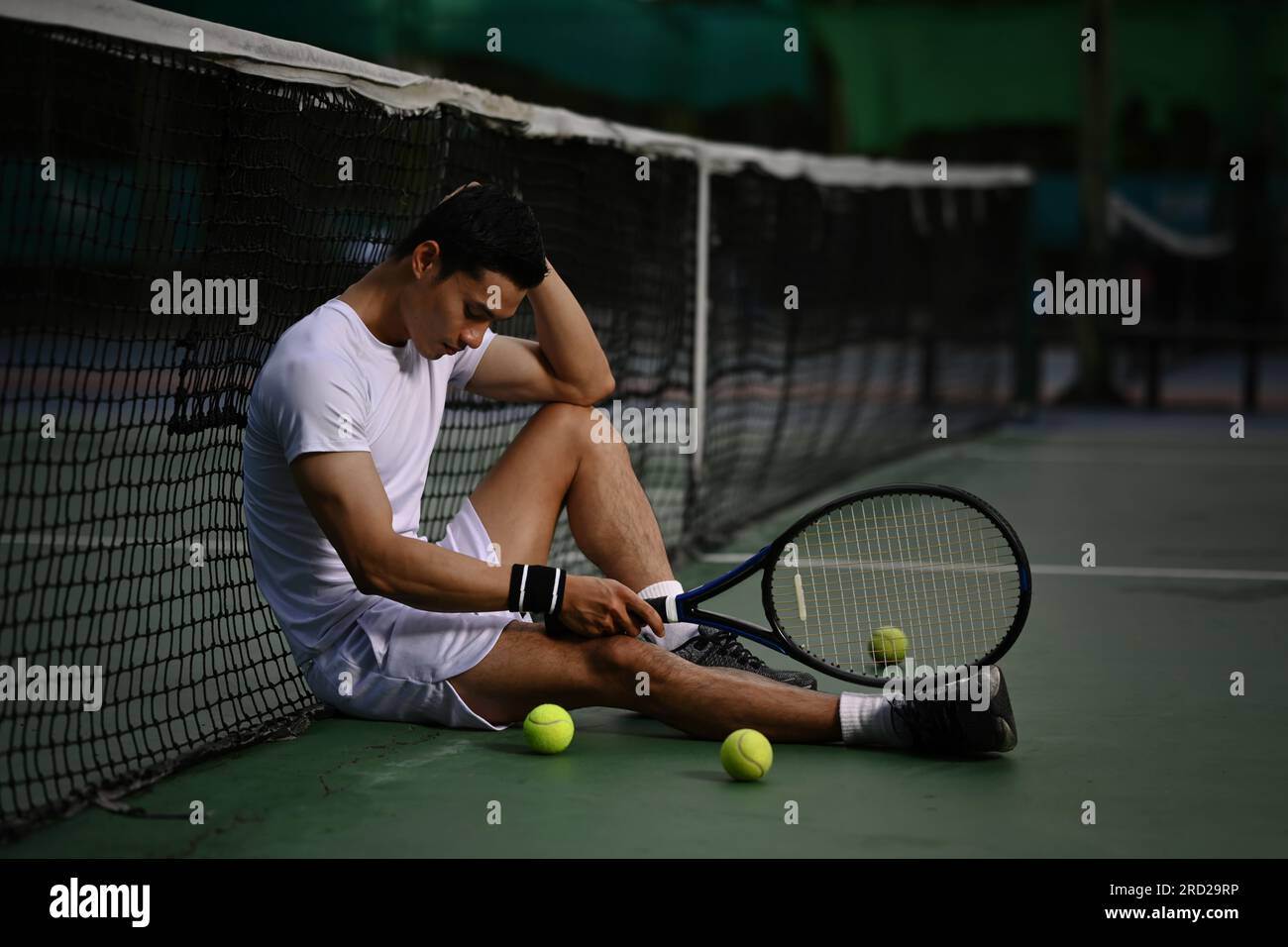 Giocatore asiatico di tennis maschile deluso seduto sul campo, turbato per la perdita di concorrenza, emozioni di fallimento Foto Stock