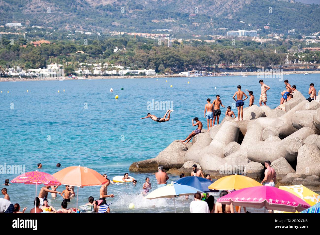 Un giovane si tuffa in mare dalle rocce vicine per rinfrescarsi in una calda giornata estiva. Altri si mettono in fila per seguirlo in mare. Marbella, Spagna Foto Stock