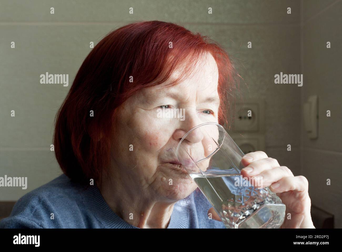 donna anziana che beve acqua minerale dal bicchiere. Ritratto in primo piano di una donna anziana dai capelli rossi in maglione blu, dissetante, a casa. Acqua per guarigione Foto Stock