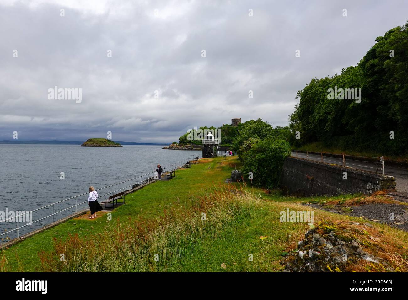 Persone che guardano il passo a Oban Bay con il faro e il castello di Dunollie sullo sfondo, Oban, Scozia, Regno Unito. Foto Stock