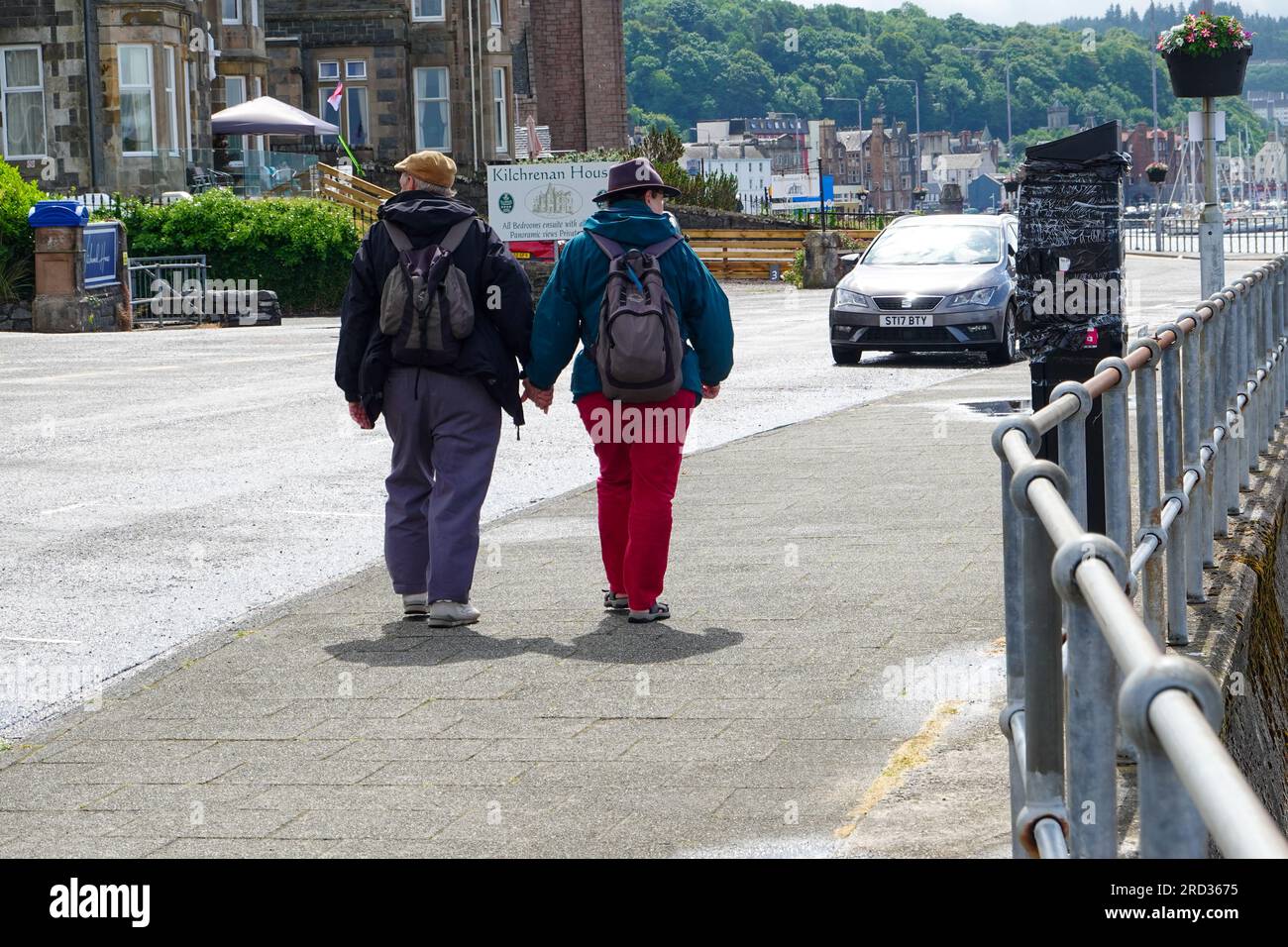Coppia matura, che indossa cappelli e zaini, cammina lungo la spianata, tenendosi per mano, Oban, Scozia, Regno Unito. Foto Stock