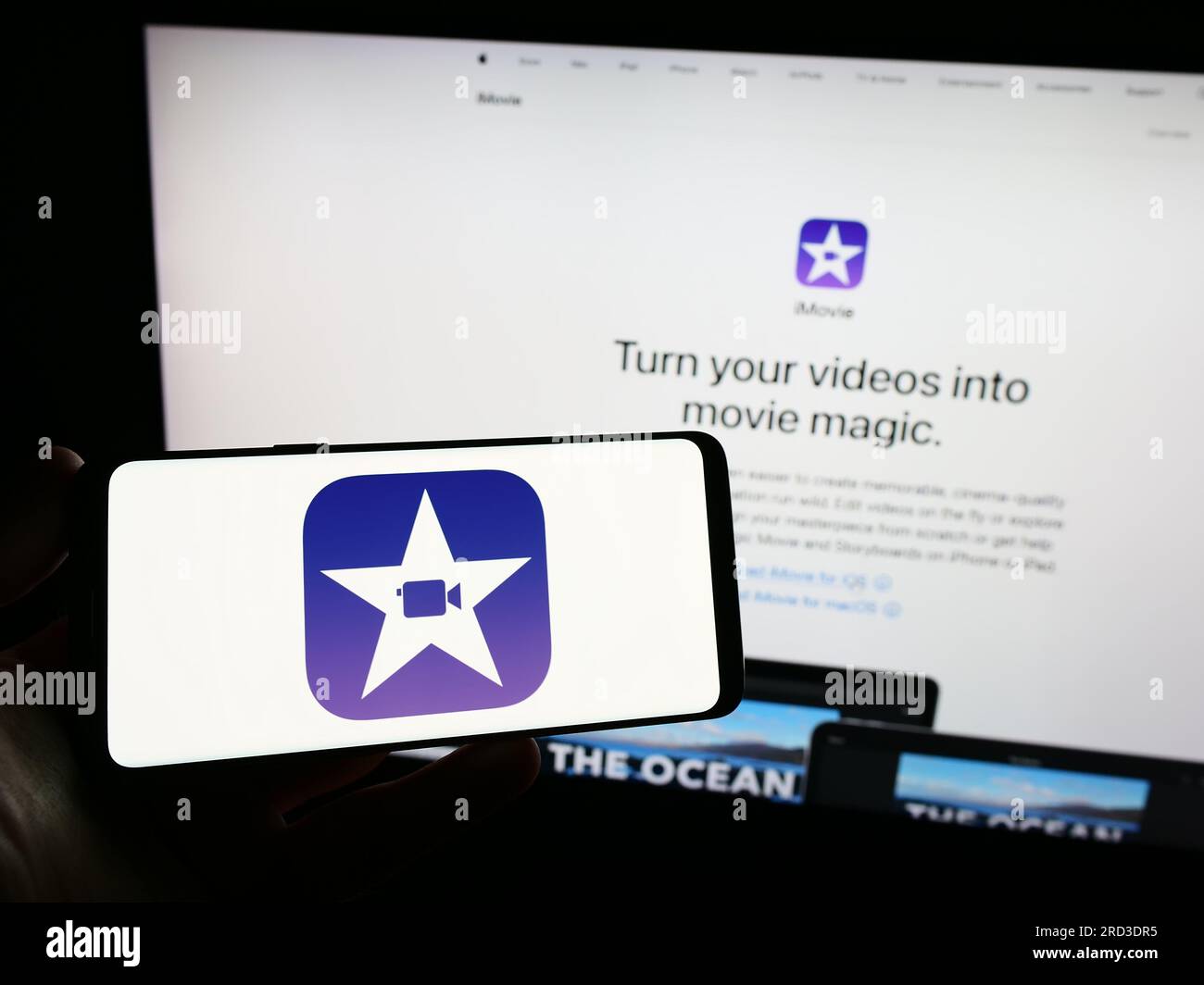 Persona che possiede uno smartphone con il logo del software di editing video iMovie (Apple Inc.) sullo schermo davanti al sito Web. Concentrarsi sul display del telefono. Foto Stock
