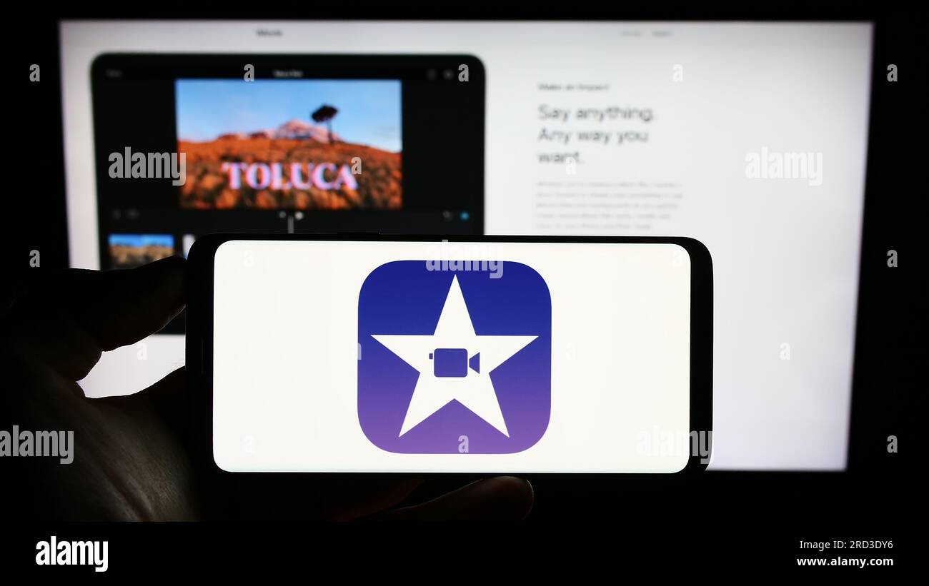 Persona che possiede il cellulare con il logo del software di editing video iMovie (Apple Inc.) sullo schermo davanti alla pagina Web. Concentrarsi sul display del telefono. Foto Stock