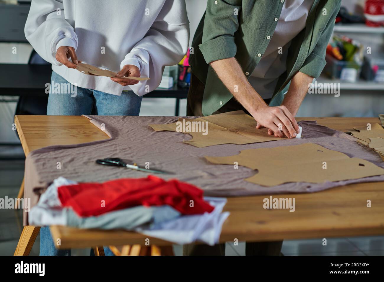 Vista ritagliata di un'artigiana afroamericana che tiene un modello di cucito mentre un collega disegna il tessuto sul tavolo in uno studio di stampa, collaborativo Foto Stock