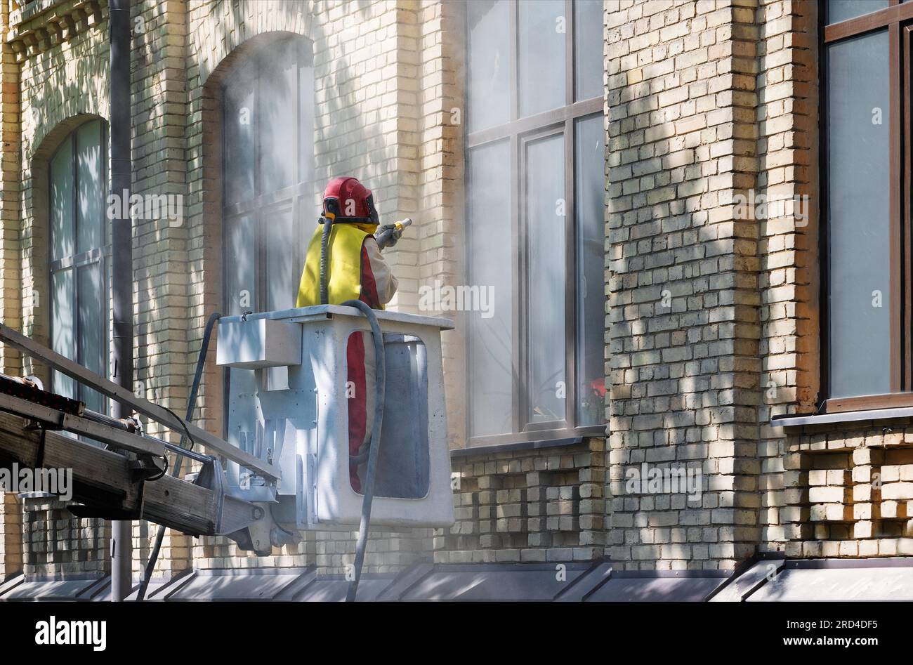 Un lavoratore in tuta protettiva pulisce la facciata di una vecchia casa in mattoni con una sabbiatrice industriale. Foto Stock