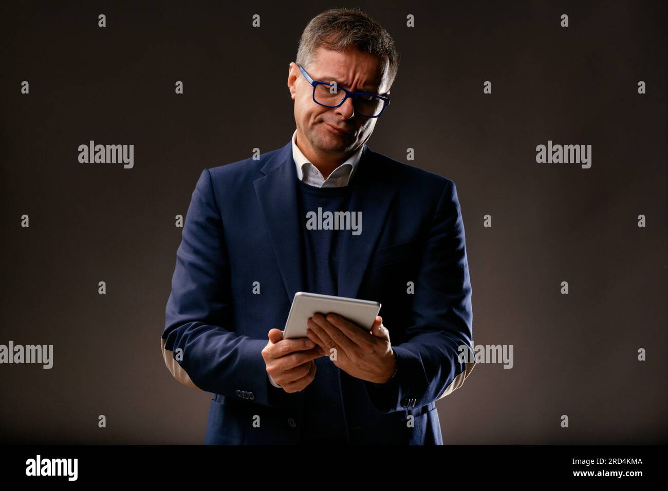 Un elegante uomo di mezza età mostra scetticismo mentre guarda lo schermo di un tablet. Indossare giacca, camicia e occhiali, leggere notizie cattive o deludenti, sighin Foto Stock