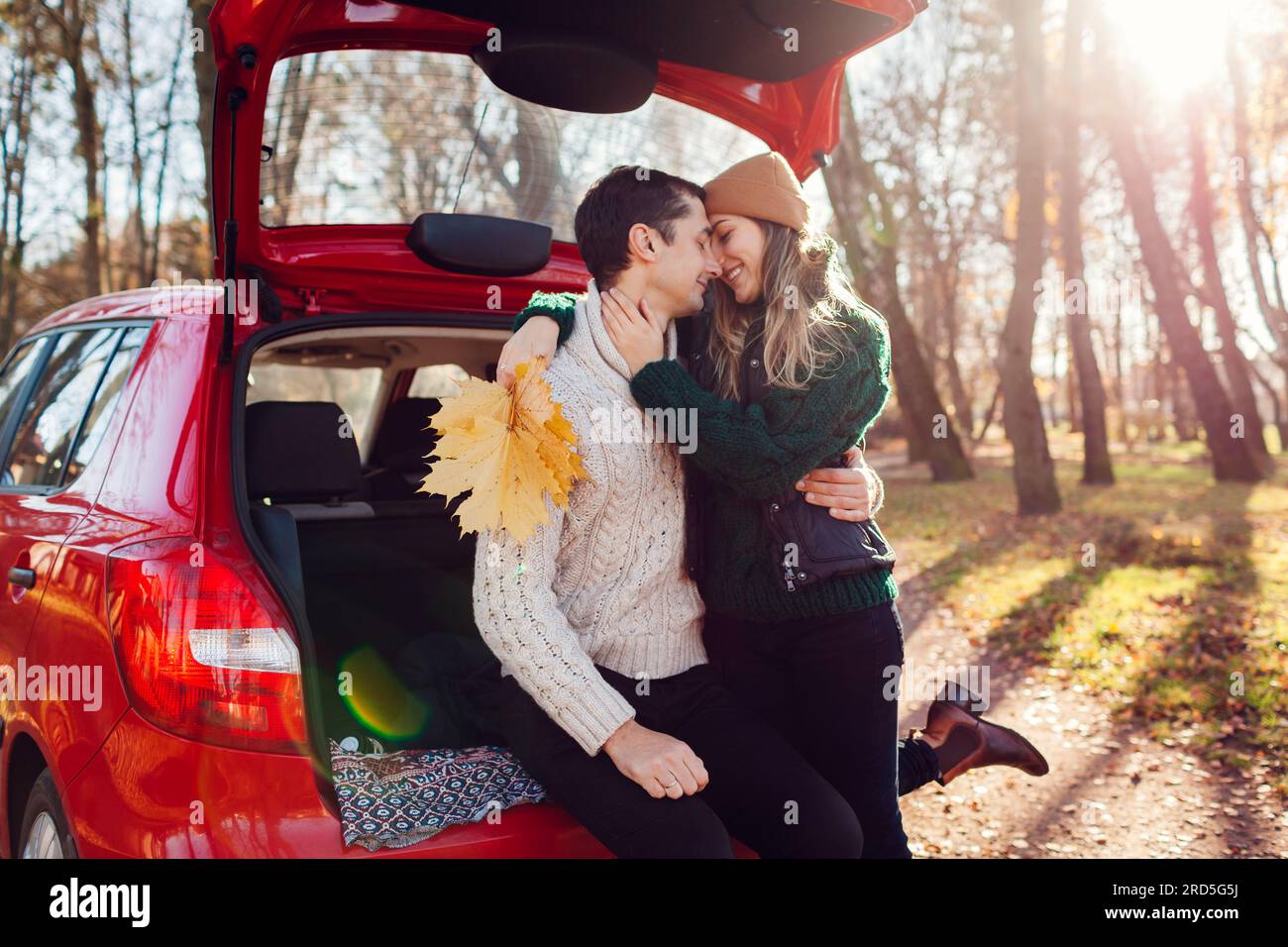 Coppia felice che ama gli appuntamenti nel bagagliaio dell'auto che si abbraccia nella foresta autunnale. Marito e moglie viaggiano in auto. Giovane donna con bouquet di giallo raccolto Foto Stock