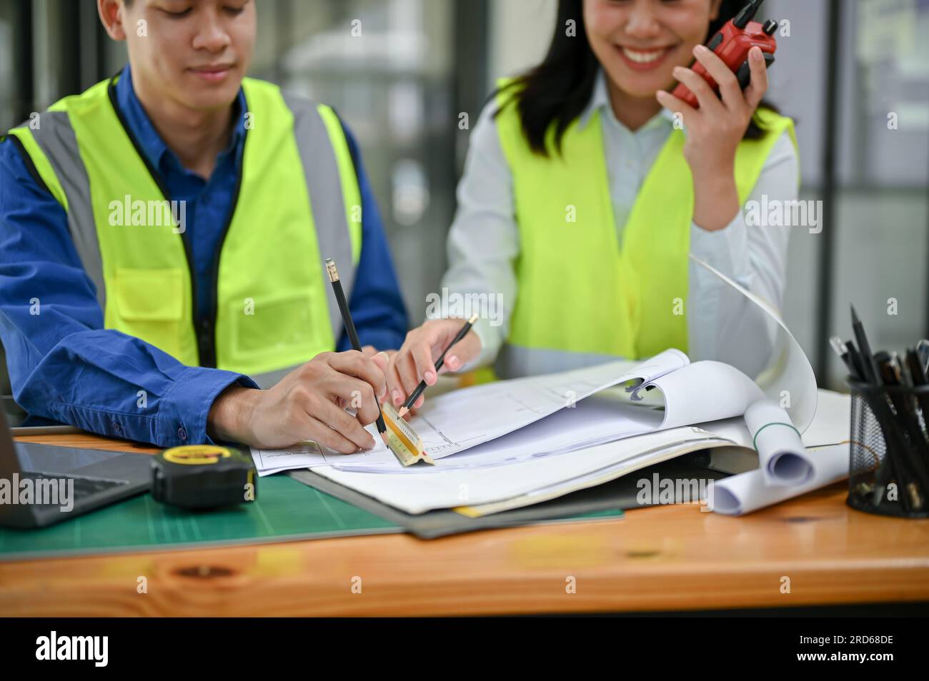 Due ingegneri edili asiatici professionisti che indossano giubbotti di sicurezza stanno controllando un progetto, discutendo del lavoro e lavorando insieme in ufficio. Foto Stock